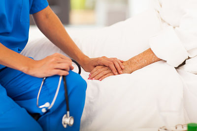 Picture of a nurse in scrubs (no face to show) sitting on the edge of a patients bed with one hand holding a stethoscope and the other touching older female patients hand (no face to show)