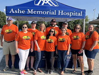 Picture of 10 (males and females) Ashe Memorial Hospital Volunteers standing outside under a Ashe Memorial Hospital tent wearing matching t-shirts.
