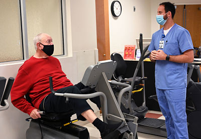 Picture of Marc Boston, Clinical Exercise Physiologist for Ashe Memorial Hospital, standing in A Cardiopulmonary Rehabilitation Program Center Room wearing scrubs and a mask looking at an older male patient who is sitting down using exercise machinery