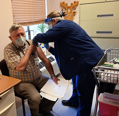 Picture of an older male patient wearing a mask and getting a shot by a Nurse that is wearing gloves, a face mask, and face shield