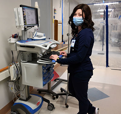 Picture of The Director of Inpatient Services, Valeree Lentz standing in a hall way with Medical Equipment with her hands on a keyboard looking over at the camera wearing a mask and scrubs