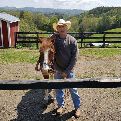 Picture of a man in the country side with mountains behind him wearing a cowboy hat standing beside a horse behind a fence at a barn