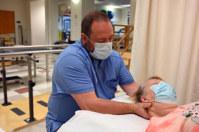 Picture of Physical Therapist Assistant, Bill Coggin. He has a female patient lying on her back. He is standing behind her with his hands under the back of her neck.