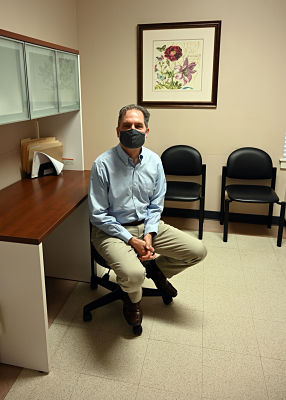 Picture of Dr. Gray sitting down on a roller chair. He is wearing a mask.