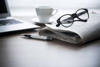 Picture of a newspaper folded with glasses on top of it, a cup of tea, a pen, and an open laptop.