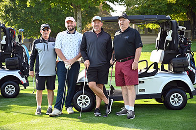 Picture of four male Golfer's standing outside on the Golf Course in front of two golf carts. They are all smiling.