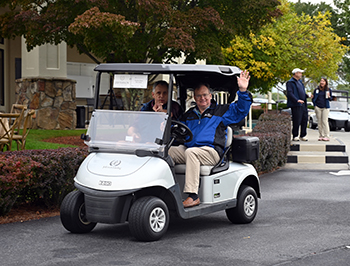 Two male golfers ride in a golf cart and wave to the audience.