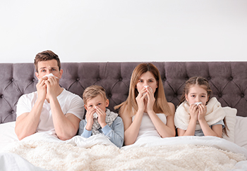 Stock photo of sick family sitting side by side in bed blowing their noses and wearing blankets.